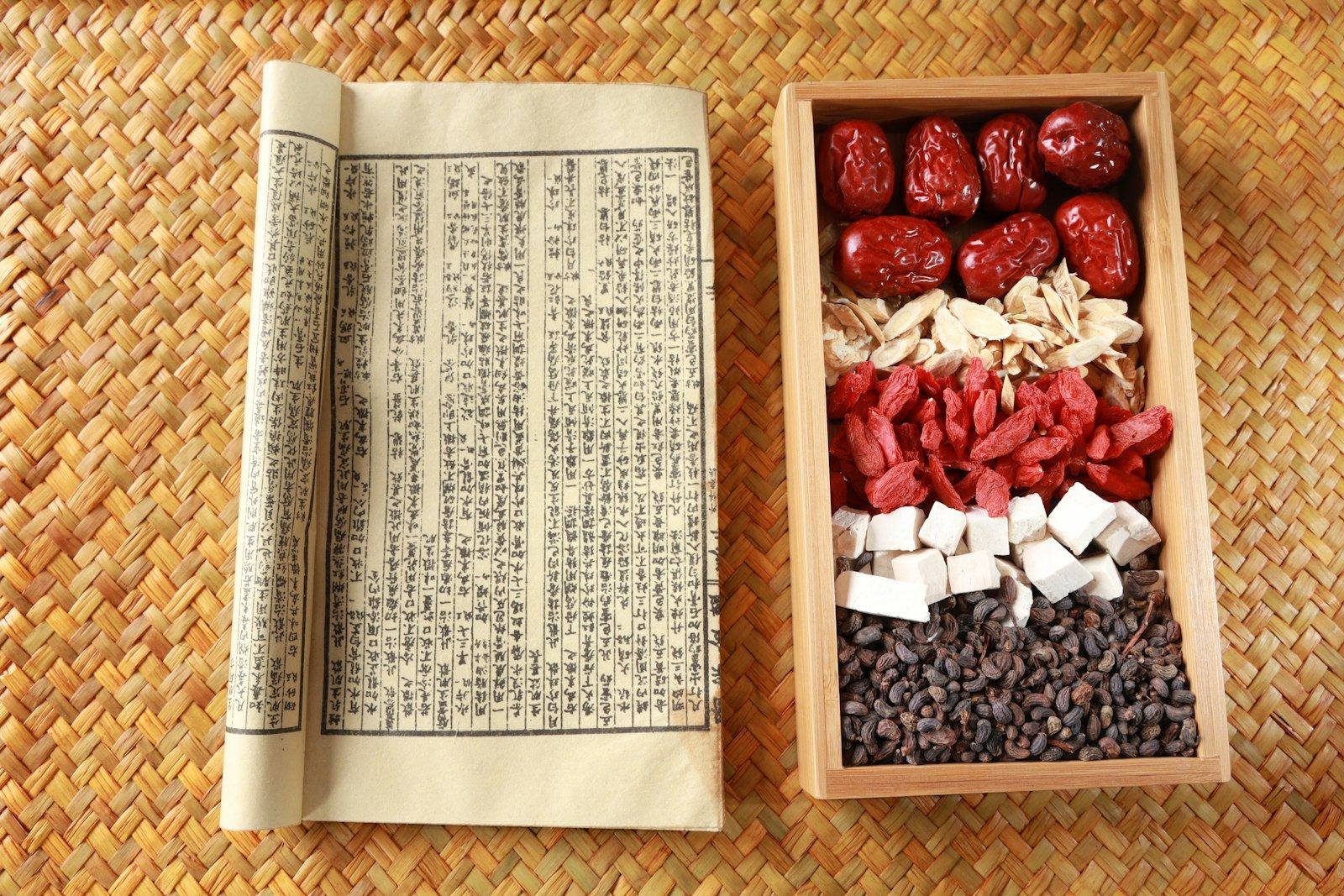A classical medical text beside a tray of jujubes, goji, and dried roots