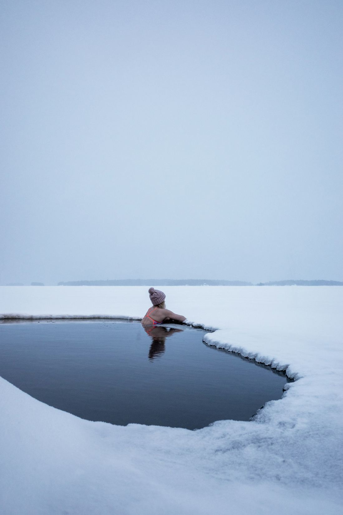 A lone swimmer in a hole cut through a frozen lake under a white sky