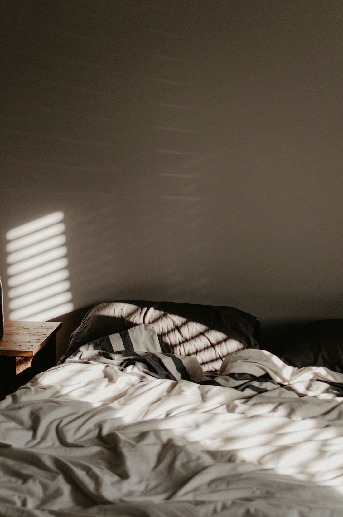 An unmade bed in a dark room, crossed by stripes of light from a blind